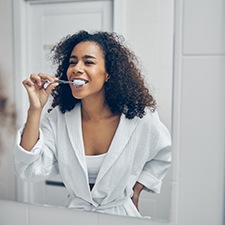 A curly-haired woman brushing her teeth in front of a bathroom mirror